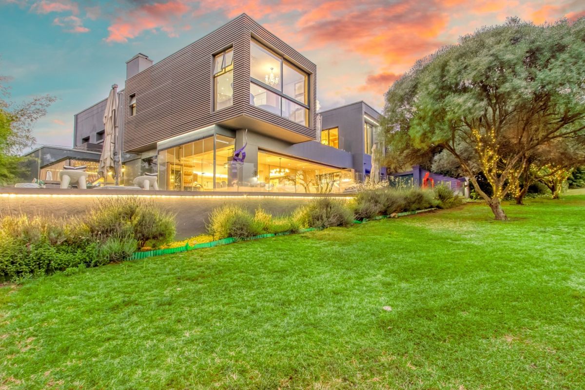 A modern architectural rear elevation featuring expansive glass walls, a landscaped garden, and integrated exterior lighting at dusk.