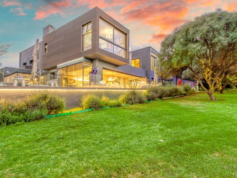 A modern architectural rear elevation featuring expansive glass walls, a landscaped garden, and integrated exterior lighting at dusk.