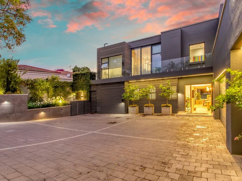 Modern two-story residence featuring a paved driveway, contemporary timber slat facade, glass balcony balustrade, and integrated exterior lighting at dusk.