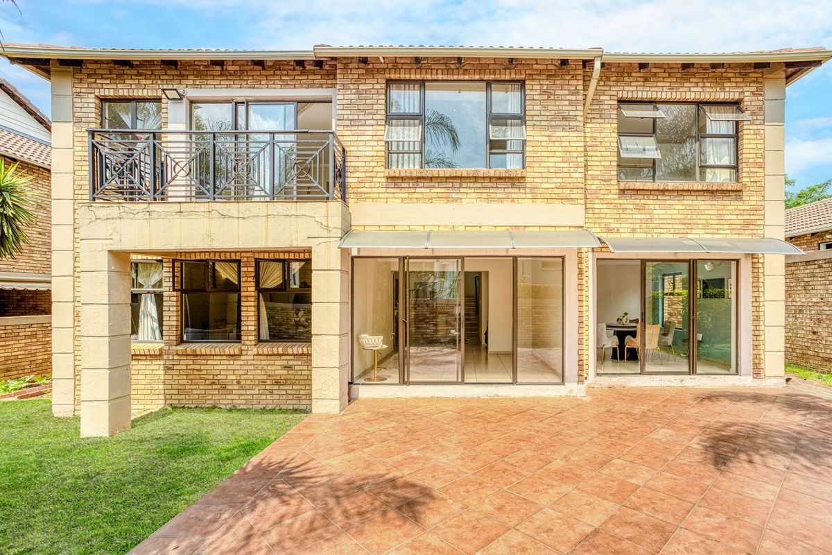 Rear view of a double-storey face brick cluster home featuring a tiled patio, balcony with metal railing, and large sliding glass doors.