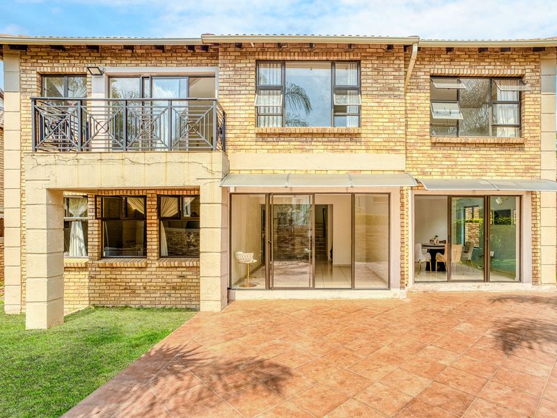 Rear view of a double-storey face brick cluster home featuring a tiled patio, balcony with metal railing, and large sliding glass doors.