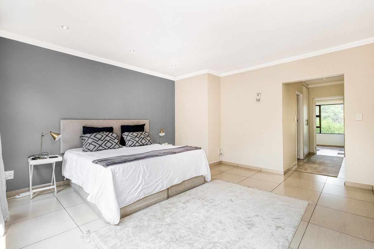 A spacious bedroom featuring neutral tiled flooring, recessed downlighting, and a feature wall, with an adjoining hallway leading to further living areas.