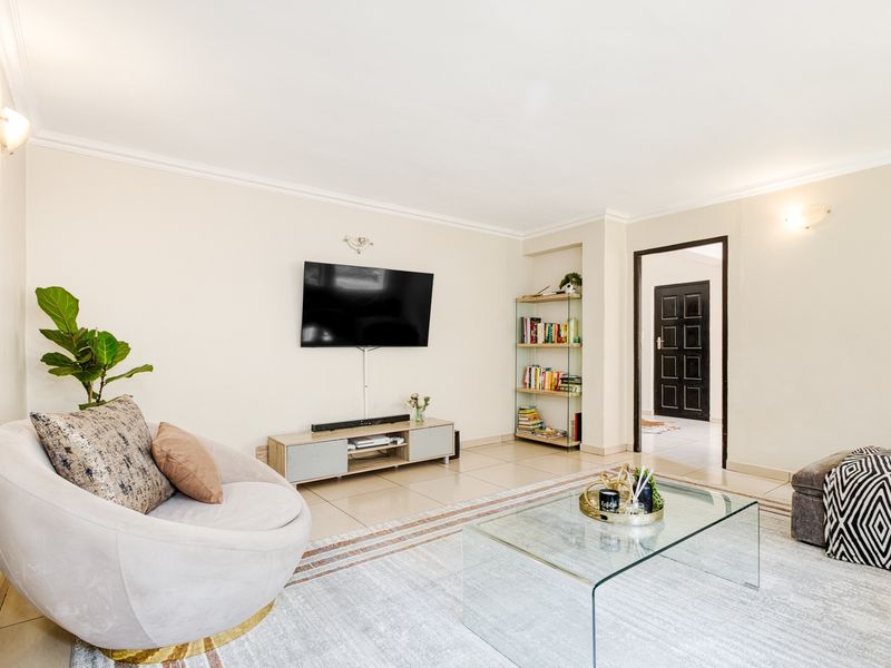 A bright and spacious living room featuring neutral tiled flooring, cove cornicing, and wall-mounted lighting, providing a comfortable and well-maintained living space.