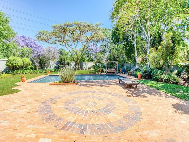 A well-maintained outdoor pool area featuring a decorative brick-paved patio, established garden landscaping, and a perimeter wall for privacy.