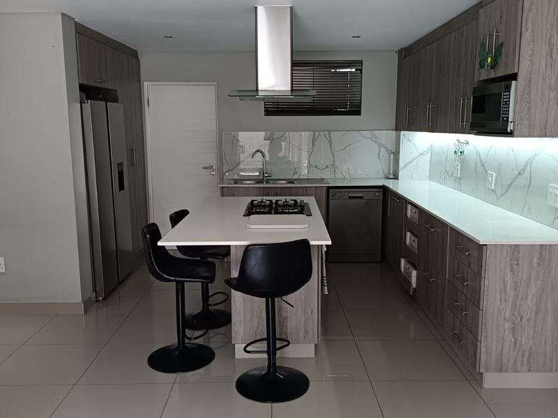 Modern kitchen featuring a central island with gas hob, stone countertops, marble-look splashback, integrated appliances, and sleek wood-finish cabinetry with under-cabinet lighting.
