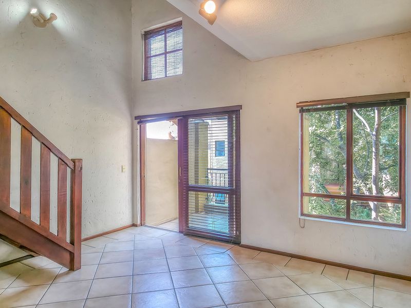 An empty living room featuring tiled flooring, a wooden staircase, vaulted ceilings, and large windows providing natural light and access to a balcony.