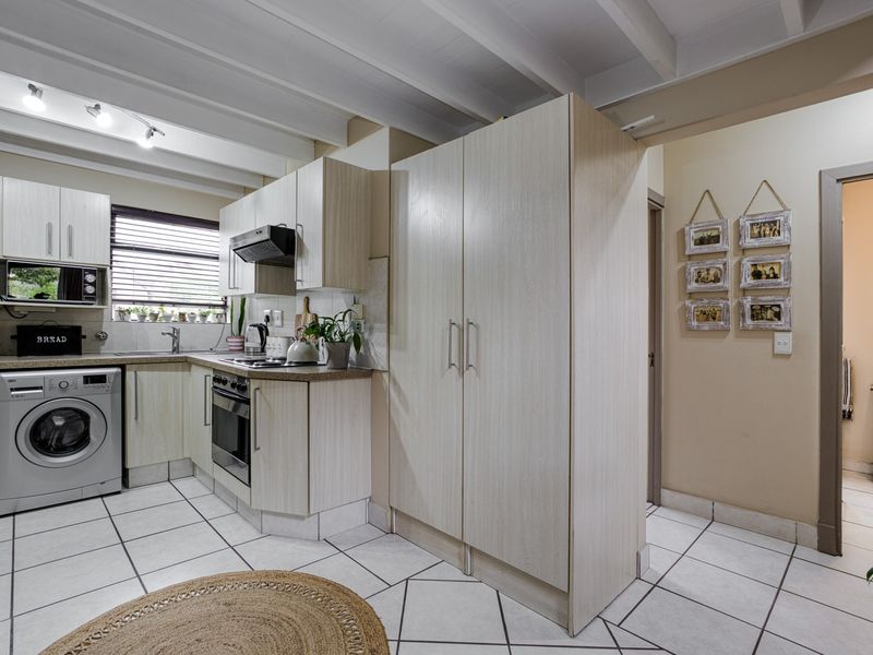 A functional kitchen featuring light-toned cabinetry, integrated appliances, tiled flooring, and an exposed beam ceiling, with an adjacent view into a bathroom area.