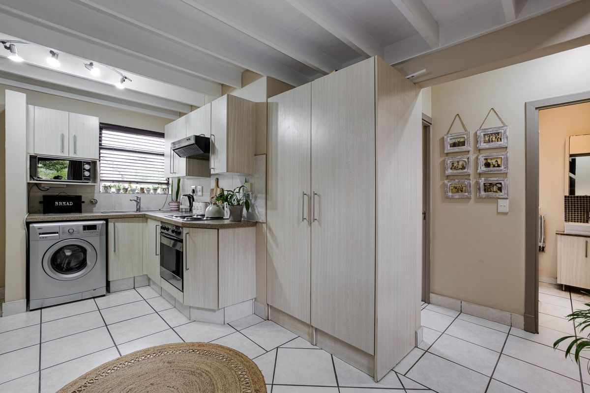 A functional kitchen featuring light-toned cabinetry, integrated appliances, tiled flooring, and an exposed beam ceiling, with an adjacent view into a bathroom area.