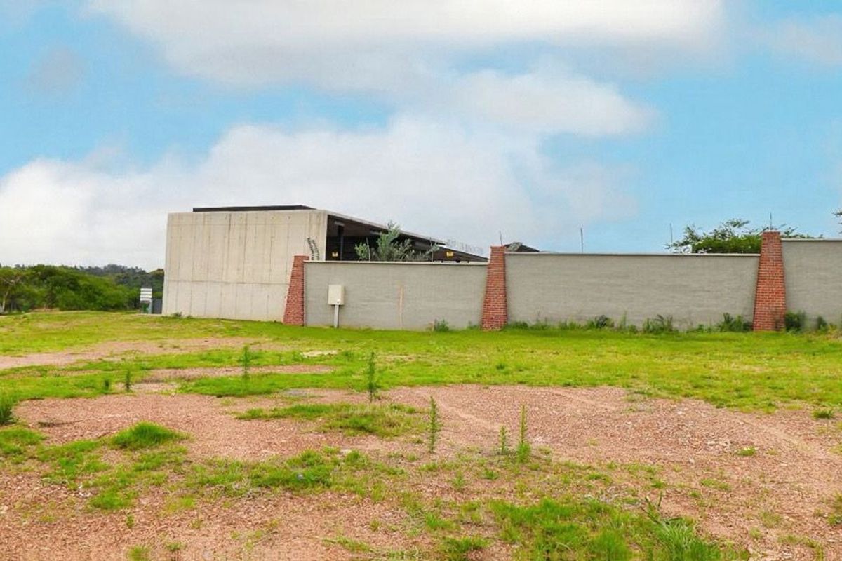 A view of a vacant residential plot featuring a modern concrete structure and a perimeter wall with brick pillars.