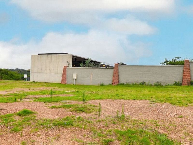 A view of a vacant residential plot featuring a modern concrete structure and a perimeter wall with brick pillars.