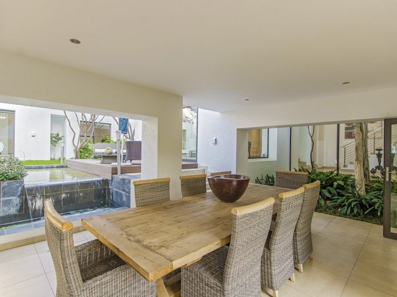 A covered outdoor patio area featuring tiled flooring, recessed lighting, and an integrated water feature overlooking a landscaped garden.