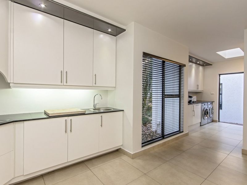 A modern, well-lit scullery featuring sleek white cabinetry, dark stone countertops, integrated sink, and tiled flooring, with a skylight and direct outdoor access.