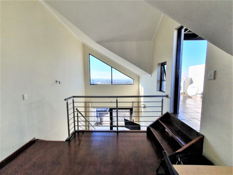 An open-plan mezzanine living area featuring hardwood floors, a modern metal balustrade, vaulted ceilings, and a unique triangular window with balcony access.