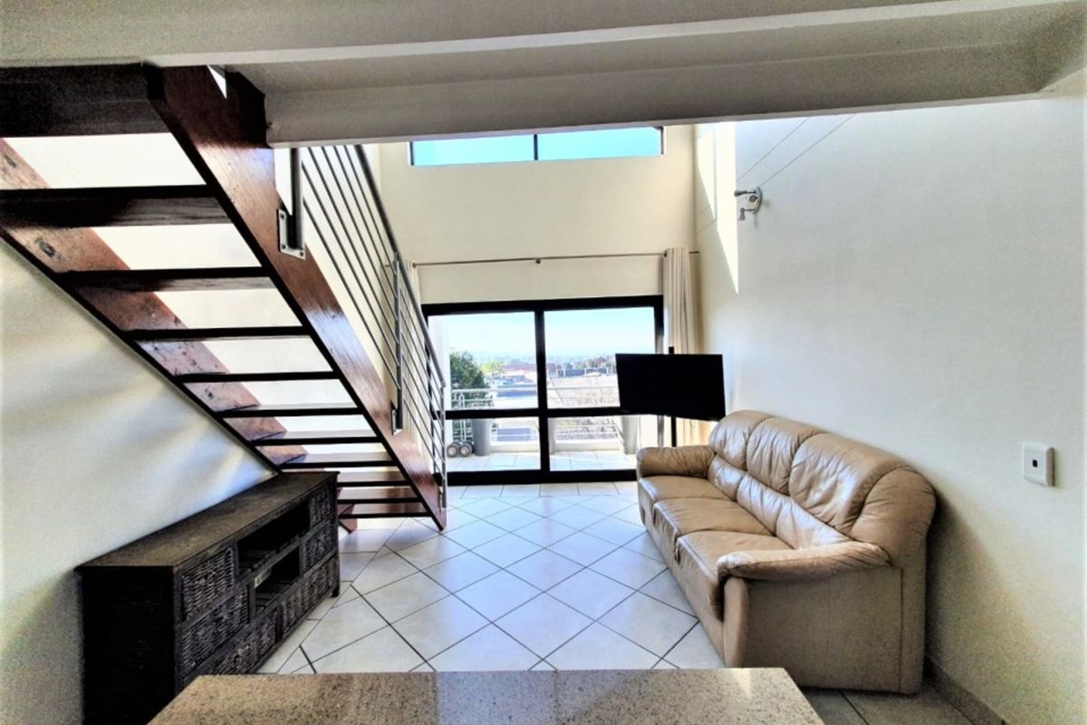 Spacious double-volume living area featuring tiled floors, an open-tread staircase with metal balustrade, and large sliding doors leading to an outdoor balcony.