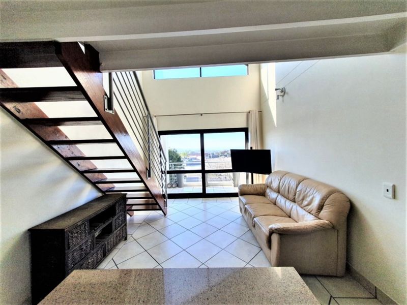 Spacious double-volume living area featuring tiled floors, an open-tread staircase with metal balustrade, and large sliding doors leading to an outdoor balcony.