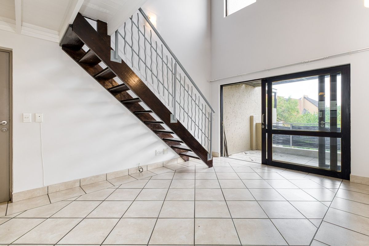 Spacious double-volume living area featuring a wooden staircase with metal balustrade, tiled flooring, and large sliding doors leading to an outdoor balcony.