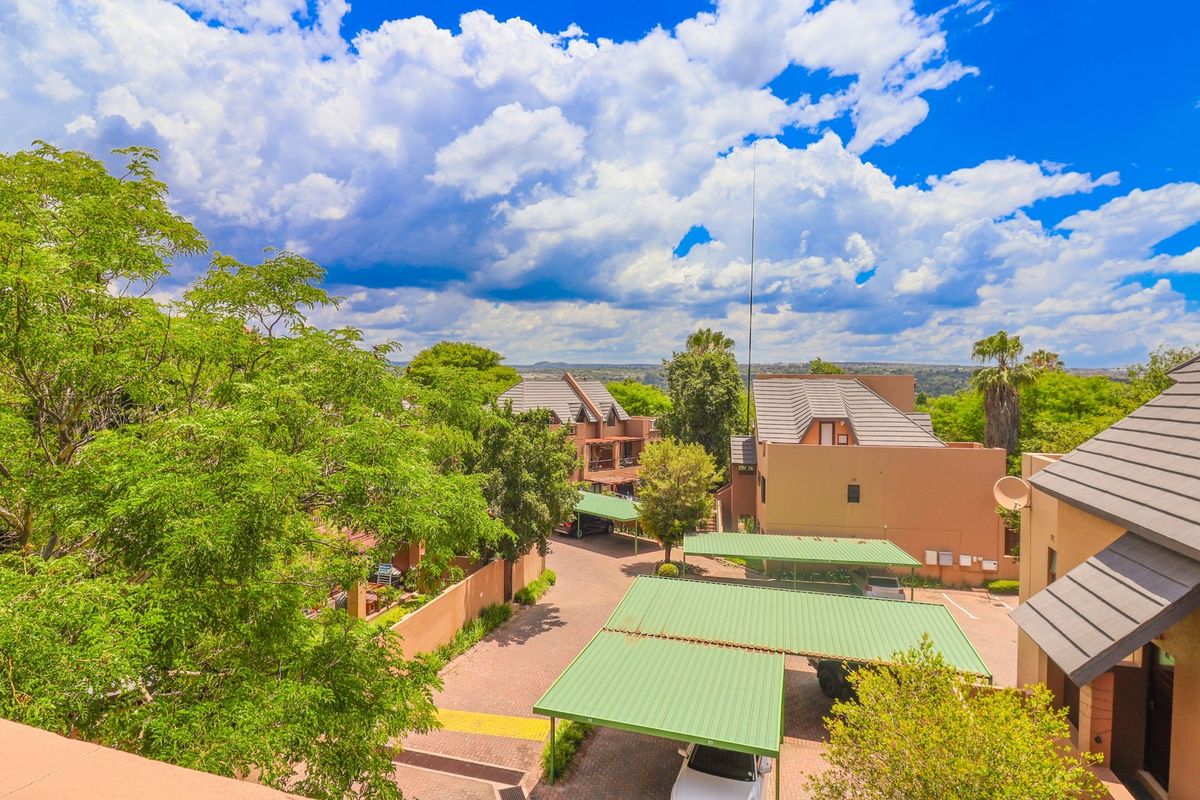 An elevated aerial view of the apartment complex grounds, featuring paved driveways, covered parking carports, and established landscaping under a bright sky.