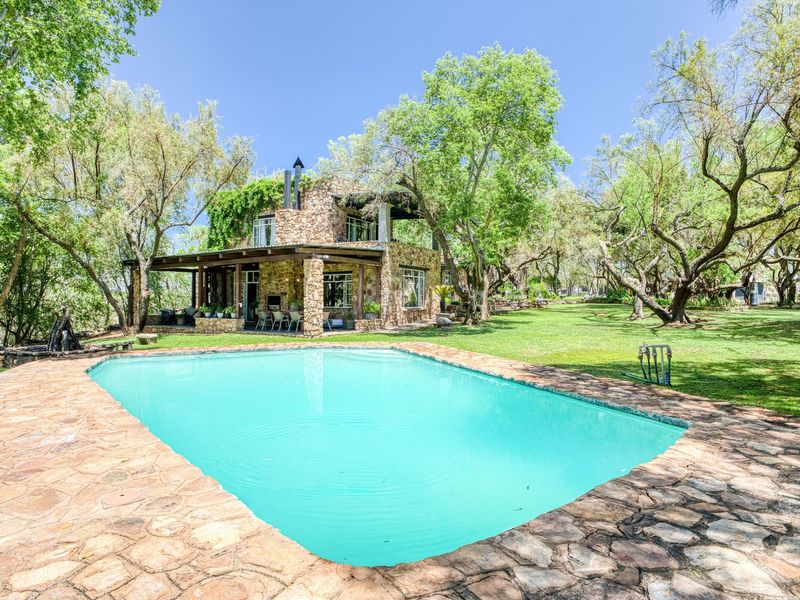 A scenic outdoor pool area featuring stone paving, a covered patio with timber beams, and a stone-clad house exterior set within a lush garden.