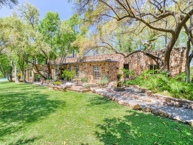 A beautiful rear garden view featuring a stone-clad house facade, a stone-paved patio, a decorative stone fountain, and a lush, well-maintained lawn with mature trees.