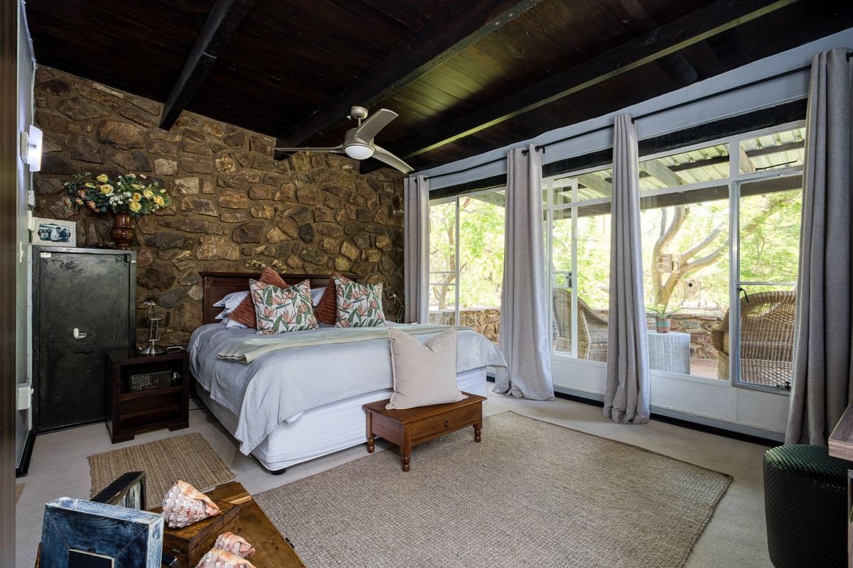 A cozy bedroom featuring a rustic exposed stone feature wall, dark timber beamed ceiling, and large sliding glass doors providing access to the patio.