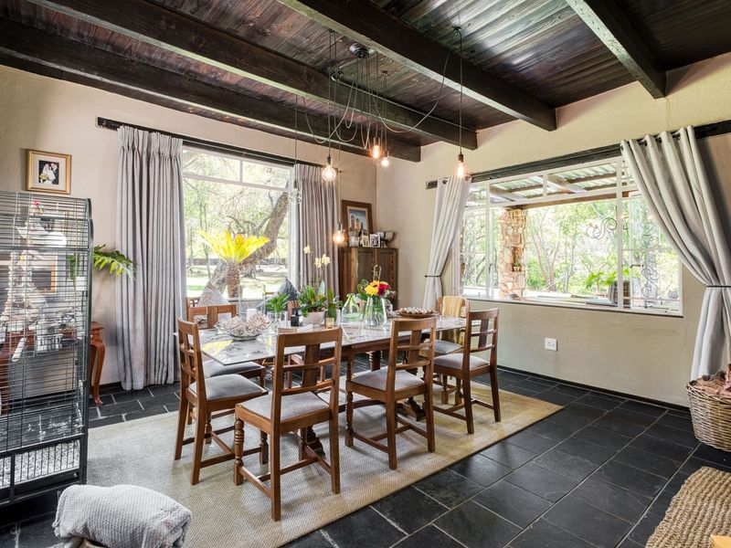 A dining room featuring exposed timber ceiling beams, dark slate tile flooring, and large windows providing natural light and garden views.