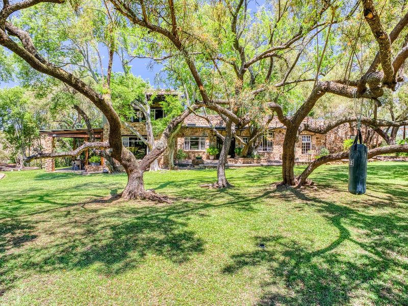 A spacious rear garden featuring a stone-clad house, a covered patio area, and mature trees providing shade over a well-maintained lawn.