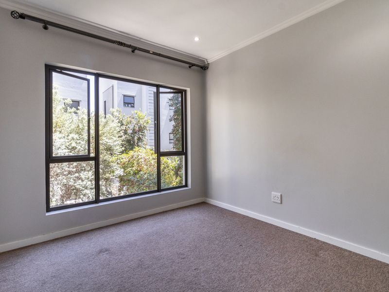 A bright, empty bedroom featuring neutral walls, carpeted flooring, and large aluminium-framed windows overlooking greenery.