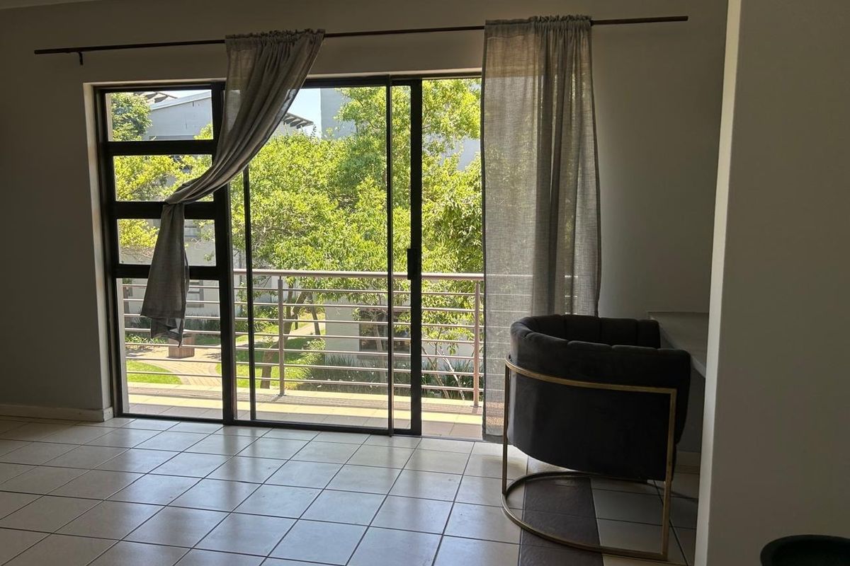 A bright living area featuring large sliding glass doors that open onto a balcony with pleasant garden views and durable tiled flooring.