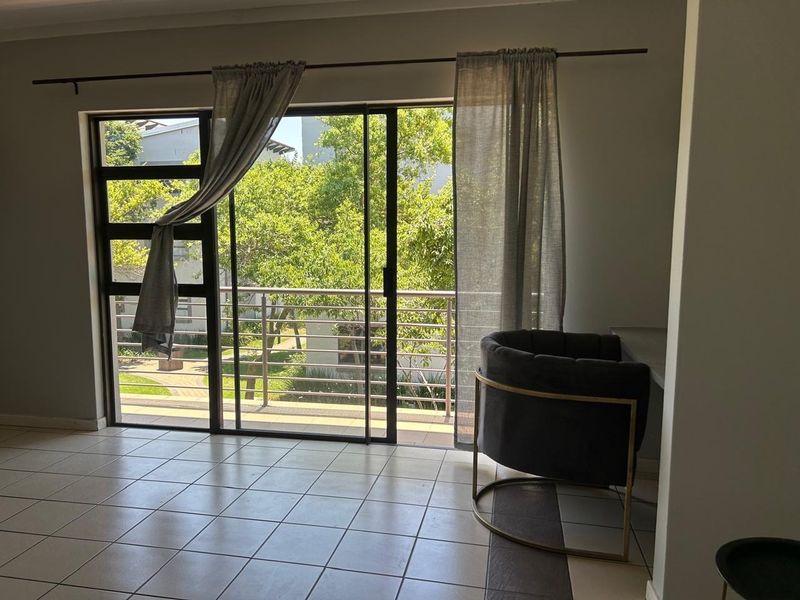 A bright living area featuring large sliding glass doors that open onto a balcony with pleasant garden views and durable tiled flooring.