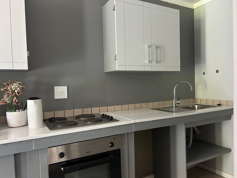 A functional kitchen area featuring a built-in oven, electric hob, stainless steel sink, and wall-mounted cabinetry with a modern grey and white finish.