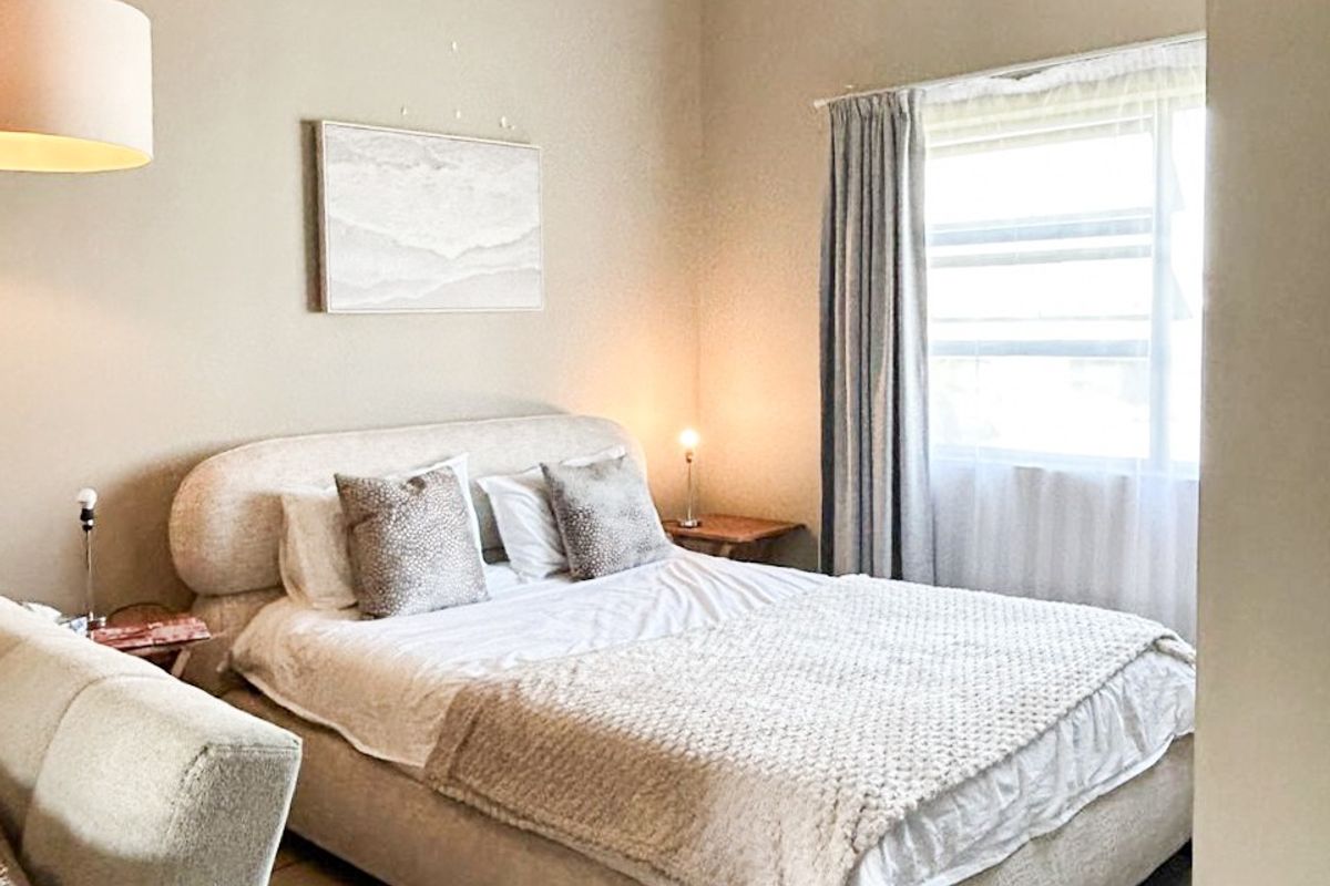 A well-lit bedroom featuring tiled flooring, exposed ceiling beams, and a window with curtains, creating a comfortable and neutral living space.