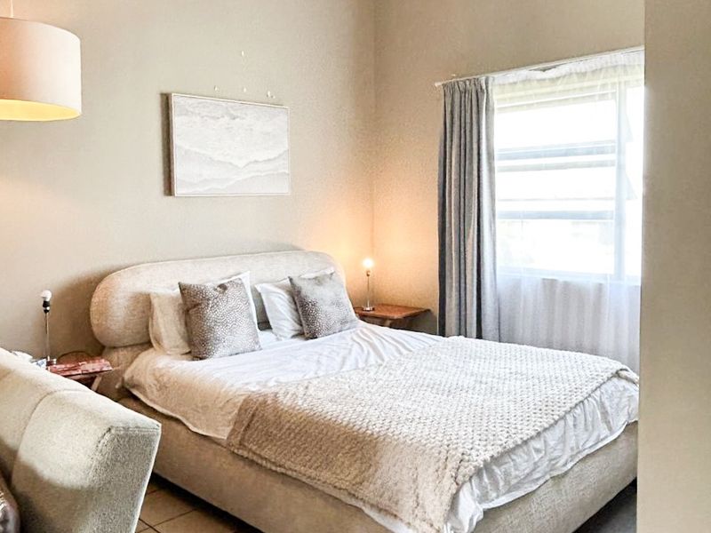 A well-lit bedroom featuring tiled flooring, exposed ceiling beams, and a window with curtains, creating a comfortable and neutral living space.