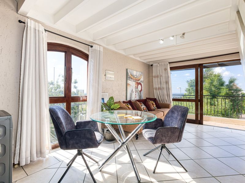 Bright living area featuring exposed beam ceilings, tiled flooring, and large windows with access to a balcony, offering natural light and outdoor views.