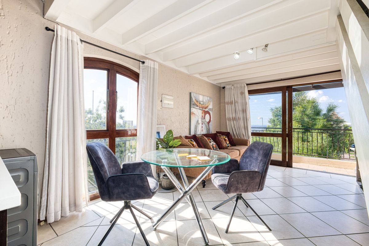 Bright living area featuring exposed beam ceilings, tiled flooring, and large windows with access to a balcony, offering natural light and outdoor views.