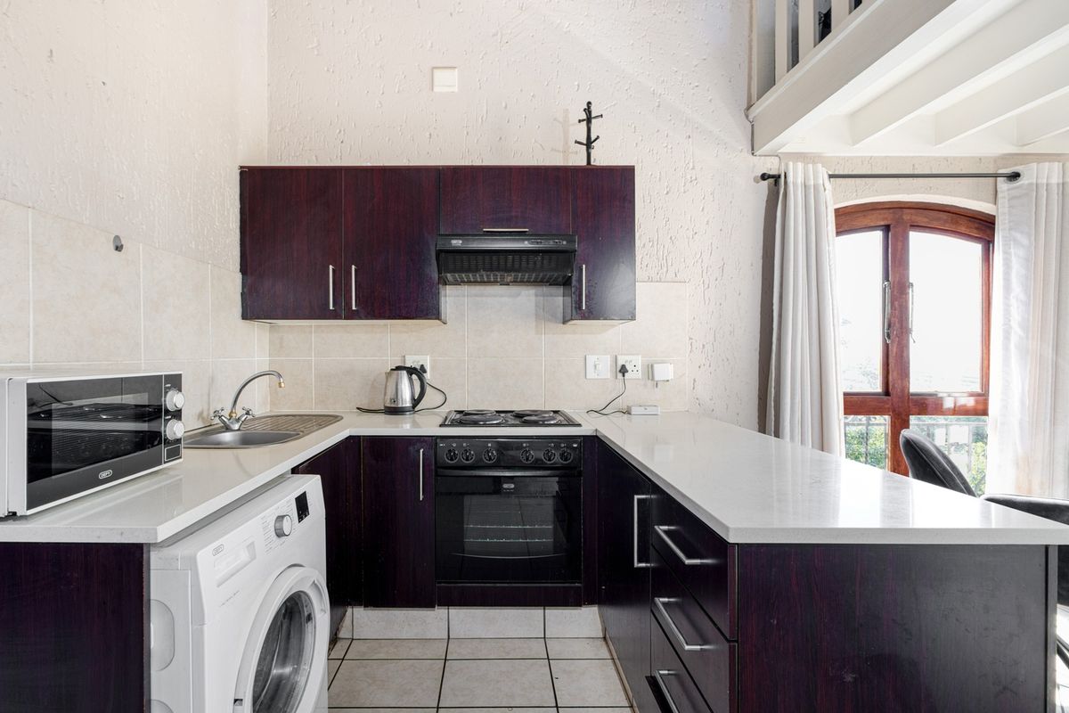 Compact kitchen featuring dark wood cabinetry, stone countertops, built-in oven, hob, and extractor fan, with an adjacent mezzanine level and natural light.