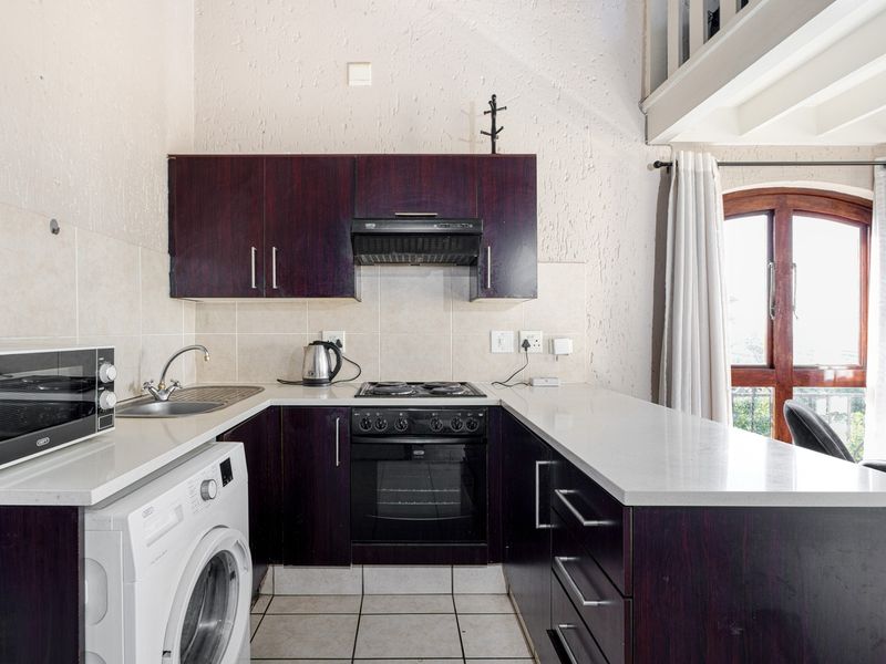 Compact kitchen featuring dark wood cabinetry, stone countertops, built-in oven, hob, and extractor fan, with an adjacent mezzanine level and natural light.