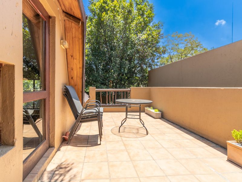 A sunny outdoor balcony featuring tiled flooring, a timber balustrade, and direct access to the interior through a large timber-framed glass door.