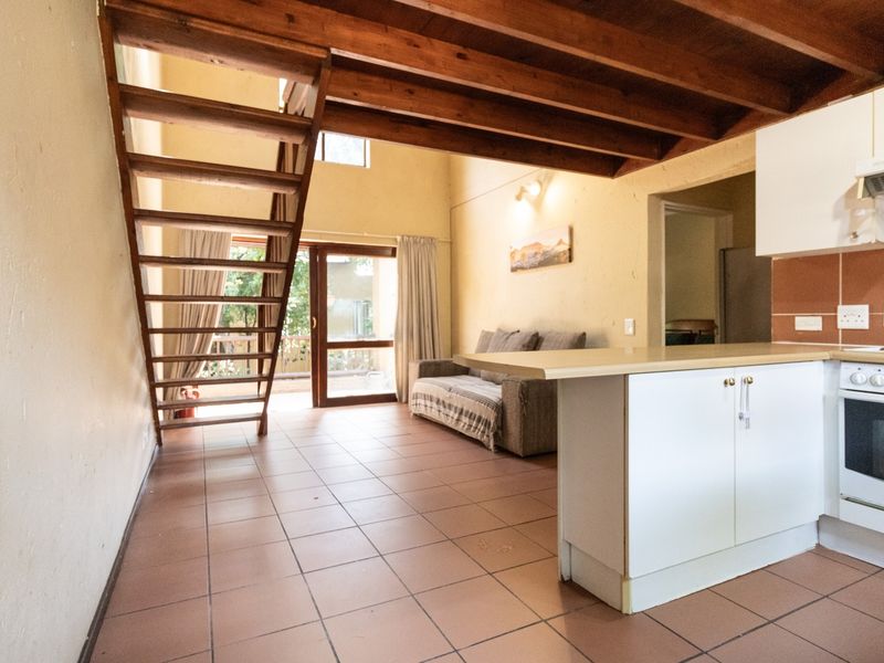 Open-plan living area featuring a timber staircase, exposed ceiling beams, tiled flooring, and a functional kitchen with built-in appliances and cabinetry.