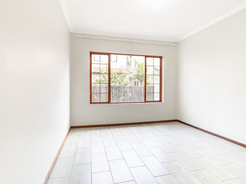 A bright, empty bedroom featuring neutral tiled flooring, clean white walls, and a large wooden-framed window providing natural light.