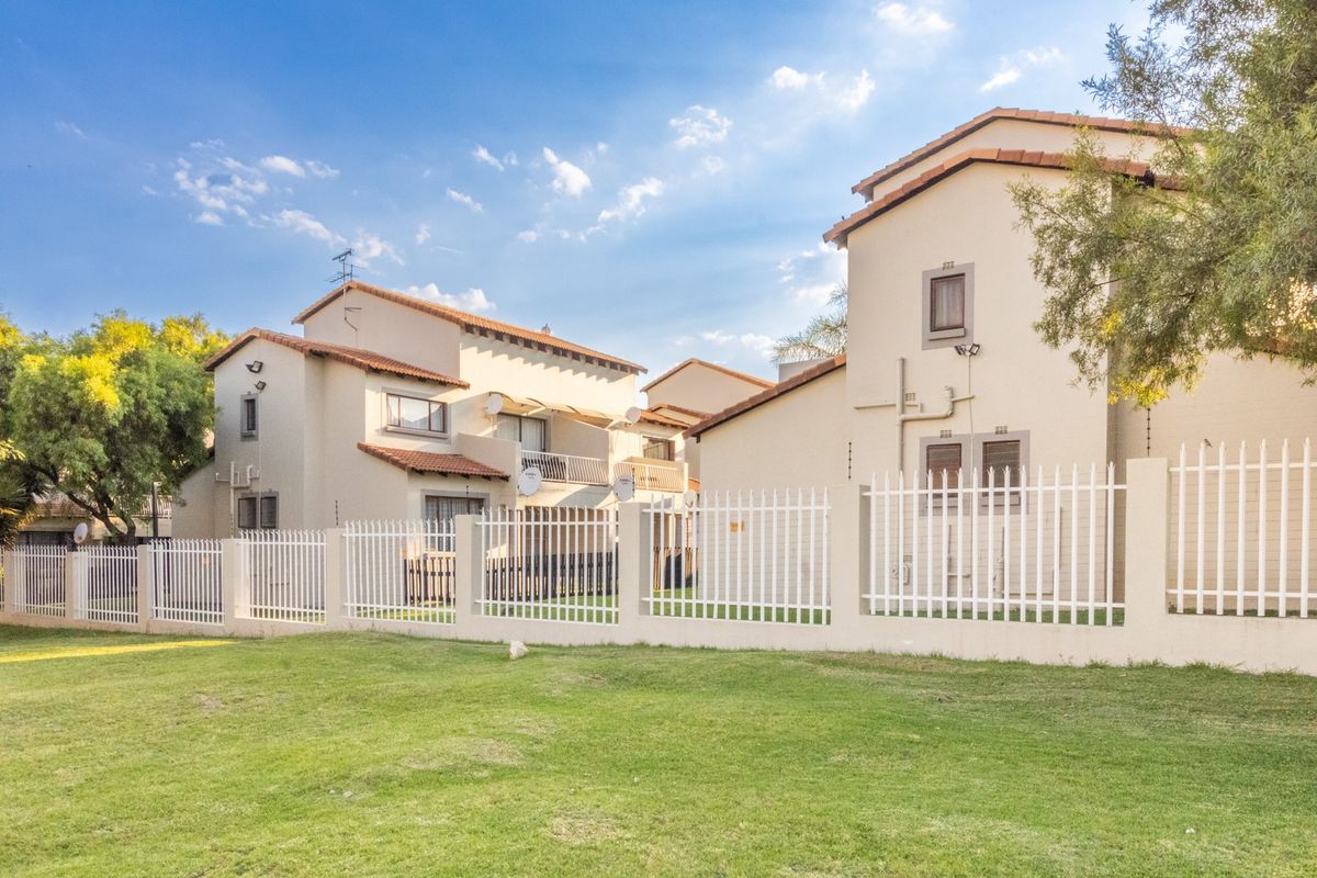 Rear view of townhouse complex featuring multi-story units with tiled roofs, balconies, and secure perimeter fencing set against a well-maintained lawn.