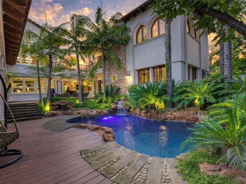 A beautifully landscaped backyard featuring a custom rock-edged swimming pool with a water feature, surrounded by timber decking and lush tropical greenery.