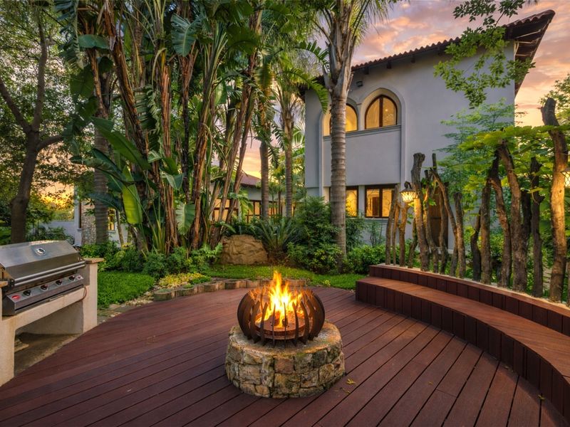 A beautifully landscaped outdoor entertainment area featuring a timber deck, built-in barbecue, and a stone fire pit, set against a lush garden backdrop.
