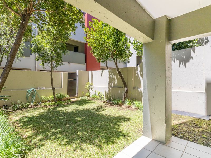 A private ground floor garden area featuring a small lawn, mature trees, and a covered tiled patio space with concrete structural pillars.