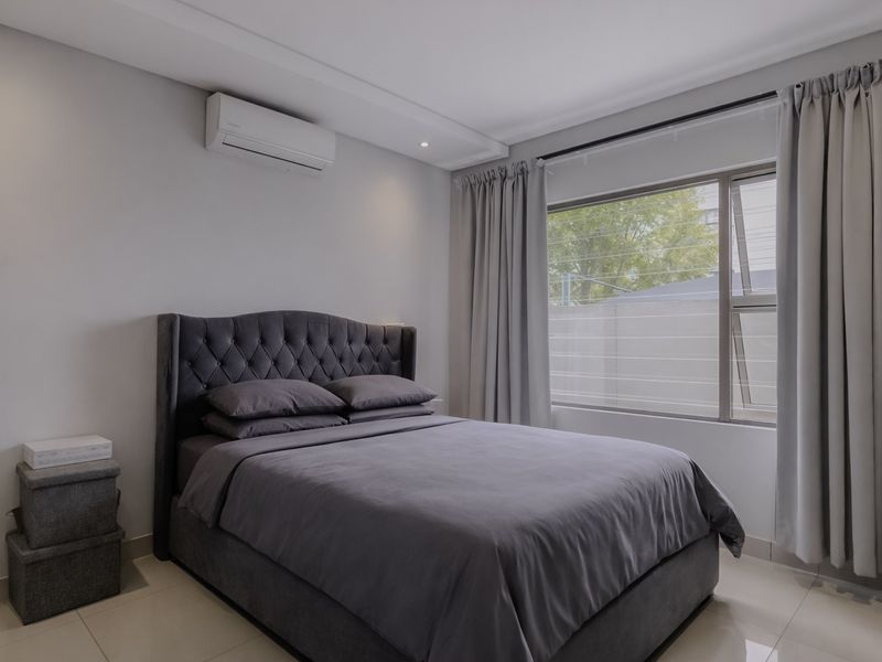 A well-maintained bedroom featuring neutral wall tones, tiled flooring, an integrated air conditioning unit, and large windows with modern curtain tracks.