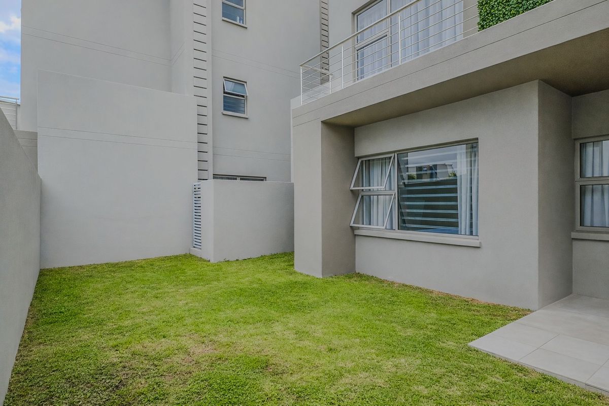 A well-maintained rear exterior view of the apartment featuring a private lawn, tiled patio area, and an upper-level balcony with stainless steel balustrades.