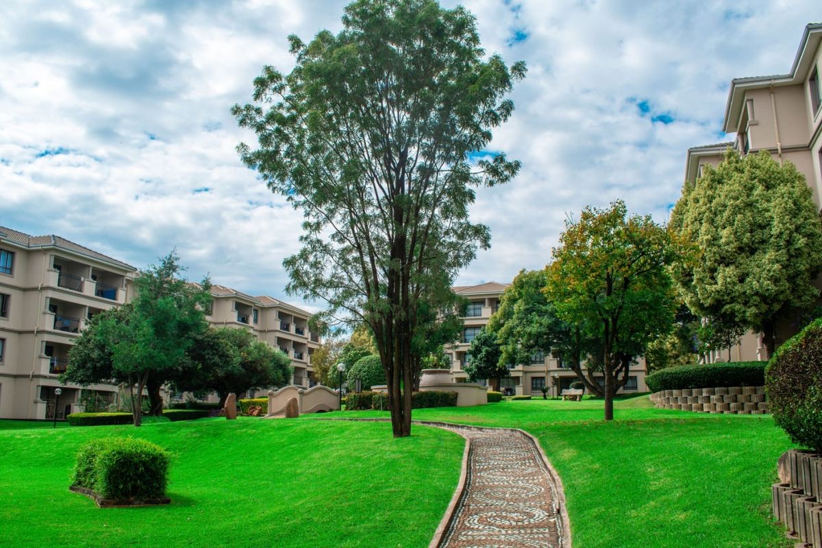 A well-maintained communal garden area featuring a stone pathway, mature trees, and manicured lawns between apartment building blocks.