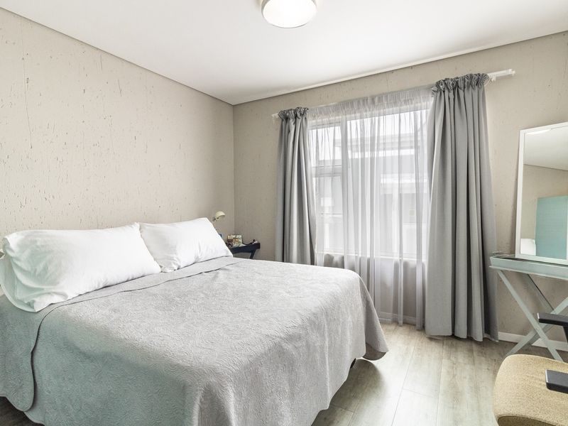 A well-lit bedroom featuring neutral textured walls, light-toned laminate flooring, and a large window with curtain tracks.