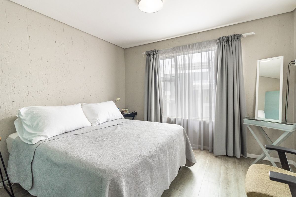 A well-lit bedroom featuring neutral textured walls, light-toned laminate flooring, and a large window with curtain tracks.