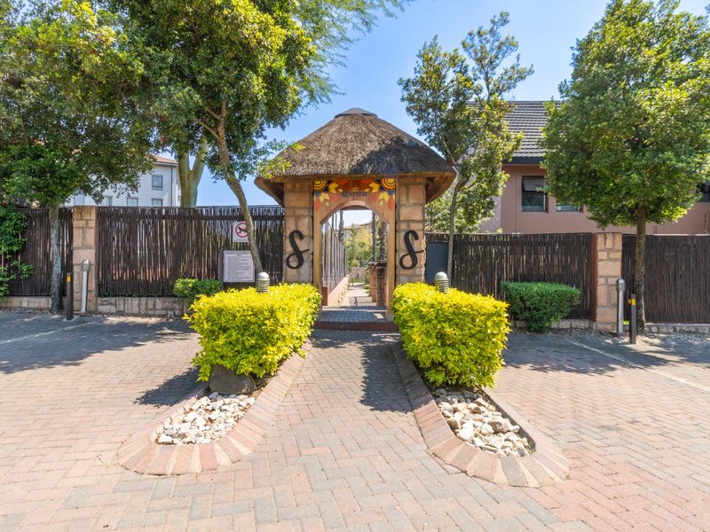 A well-maintained entrance featuring a thatched roof gateway, stone pillars, and paved driveway leading into the apartment complex.