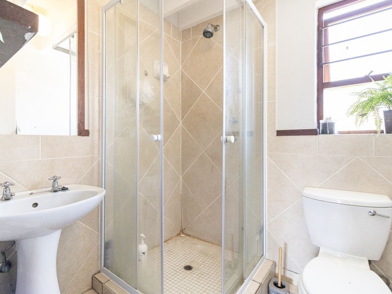 A functional bathroom featuring a pedestal washbasin, a corner shower enclosure with glass doors, and a standard toilet, finished with neutral wall and floor tiles.